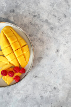 A plate with sliced mango and fresh raspberries on a textured gray surface. The mango is bright yellow with a smooth texture, while the raspberries are red and plump.