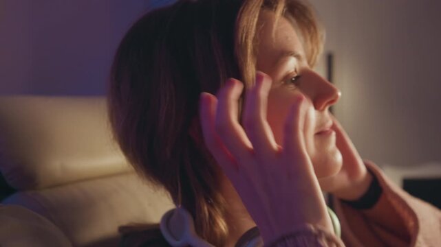 Medium close up profile of young woman seated with padded over ear headphones, hands repositioning earcups and pulling hair back as she settles into relaxed listening posture indoors