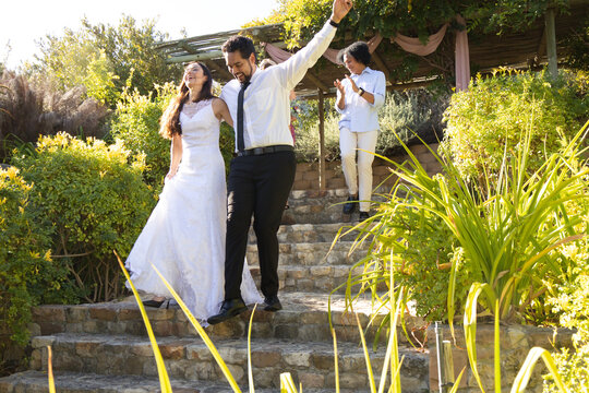 Couple descending stone stairway in garden, bride holding gown, groom raising arm showing black tie