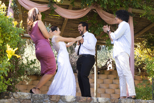 Diverse wedding party laughing while bride tugging groom's black tie under wooden pergola drapery
