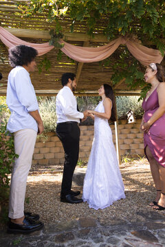 Diverse wedding party exchanging vows under pergola with pink fabric bride wearing lace gown