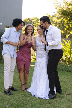 Diverse bridal party raising champagne flutes, bride wearing white lace gown, standing on lawn