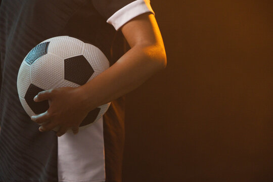 Soccer player is holding black-and-white ball against hip in studio with warm rim light, copy space