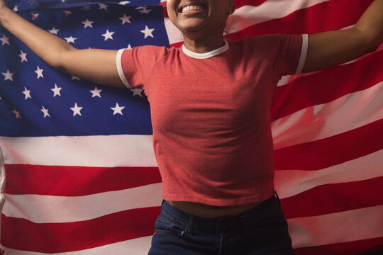 African American woman holding US flag behind, smiling in studio wearing red ringer tee, dark jeans