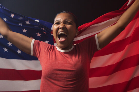 African American woman shouting and holding large American flag in studio, in red ringer T-shirt