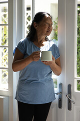 Senior Chinese woman standing by white double doors wearing light-blue shirt and pants, holding mug © wavebreak3