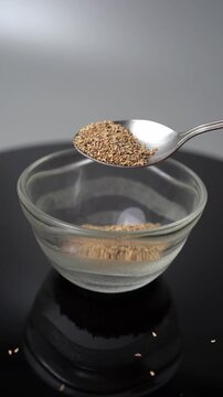 Close-up of dried Ajwain seeds (Trachyspermum ammi) falling from a spoon into a glass bowl on a dark surface