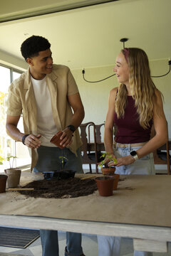 Diverse couple planting seedlings at sunroom table with terracotta pots and soil, smiling