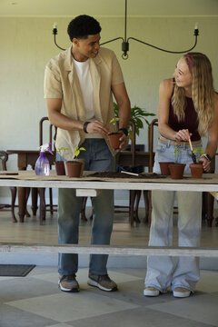 Diverse couple potting seedlings on wooden table in sunroom with terracotta pots, soil, trowel