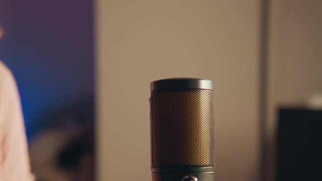 Upright condenser microphone stands in warm softly lit home studio while hand moves near grille during setup, suggesting recording, voiceover, podcast, or streaming conversation, with microphone