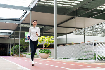 Fototapeta premium Asian athletic woman jogging on modern urban running track, holding smartphone and water bottle while wearing earphones. Healthy active lifestyle, fitness, and wellness concept.