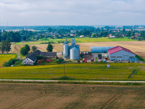 Aerial view of an agricultural farm featuring silver grain silos, large barns, and farm equipment surrounded by golden fields and green rural landscape in Jaunberze, Dobele Municipality, Latvia.