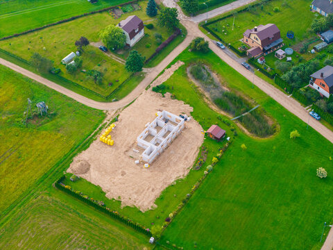 Aerial view of a residential construction site with white block walls, surrounded by green fields, rural roads, and nearby houses in Jaunberze, Dobele Municipality, Latvia.