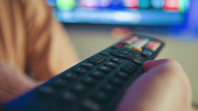 Low-angle close-up shows hand holding textured plastic remote control while thumb presses top buttons and device tilts toward blurred glowing screen, suggesting channel change, menu navigation, and