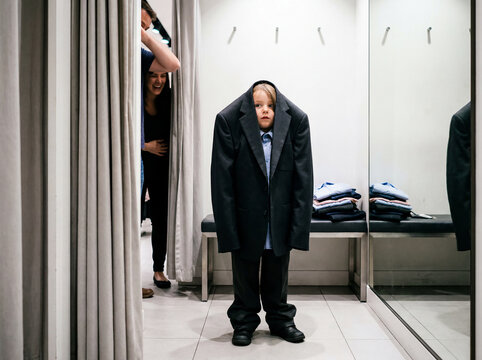 A boy in an oversized school suit is fooling around in a clothing store fitting room with his mother.