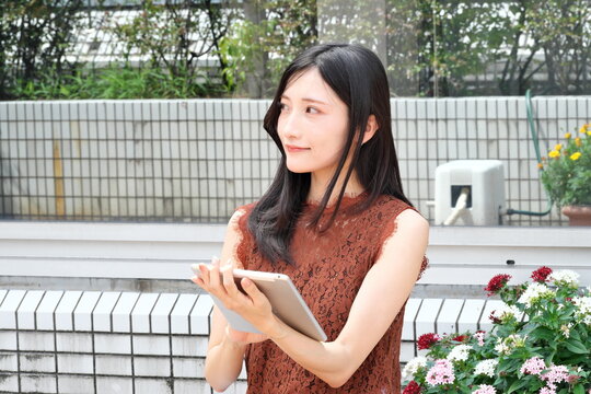 花壇のある屋外テラスでタブレットを持ち微笑む若い女性