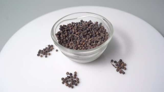 Close-up View of Dried Black Peppercorns (Piper nigrum) in a Small Glass Bowl and Scattered on a White Surface