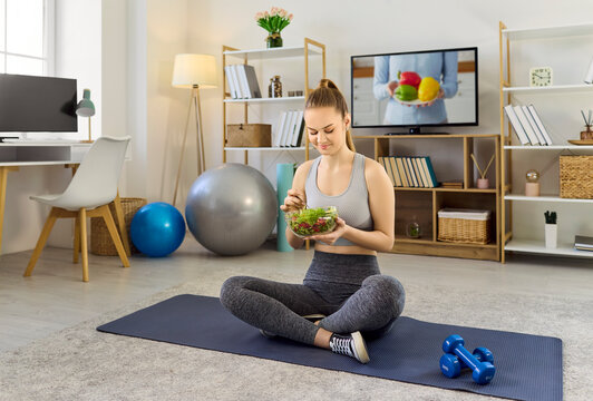 Fitness woman eating salad after home workout. She sits on a yoga mat near dumbbells, holding greens to highlight healthy nutrition and wellness at home. Concept of healthy active lifestyle