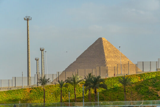 An ancient pyramid rising behind modern perimeter fencing, light towers, and a landscaped green slope