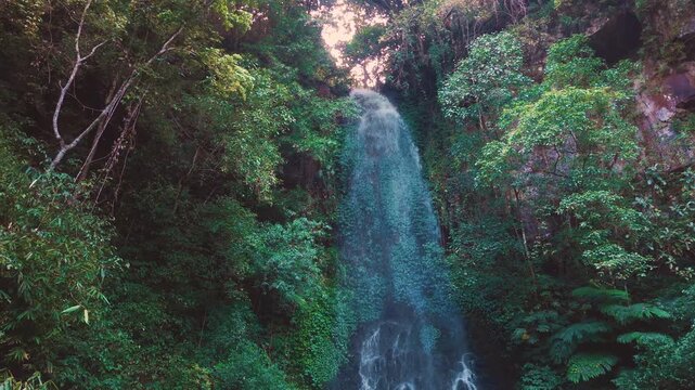 Aerial drone view of Coban Kaca waterfall flowing through dense tropical forest in Malang, East Java, Indonesia. The narrow waterfall cascades between lush green trees, creating a peaceful and seclude