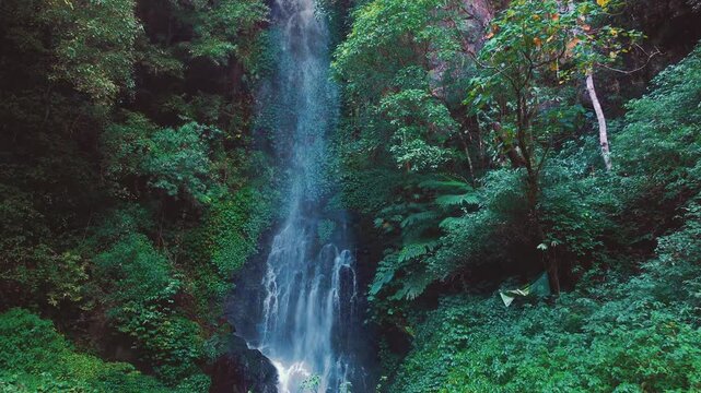 Aerial drone view of Coban Kaca waterfall flowing through dense tropical forest in Malang, East Java, Indonesia. The narrow waterfall cascades between lush green trees, creating a peaceful and seclude