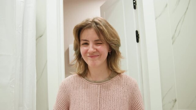 Young woman stands in bathroom doorway, moving from broad smile to playful smirk with relaxed shoulders, slight head tilt, direct gaze, warm diffuse light, calm domestic mood, with woman, smile
