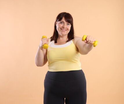 Overweight woman exercising with dumbbells on beige background. Sports equipment