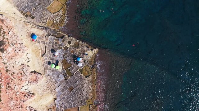 Sunbathers and swimmers relax beside ancient salt pans carved into limestone rock, viewed directly overhead by drone above the calm turquoise waters of Marsaskala, Malta.