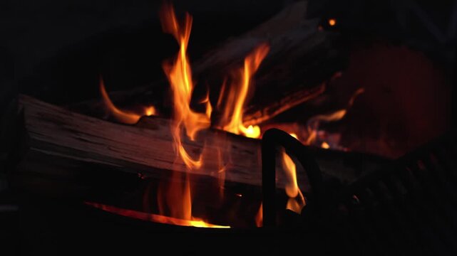 Orange campfire flames burn around stacked logs in darkness, creating a warm glowing scene with dancing fire, charred wood and deep shadows during an outdoor night gathering