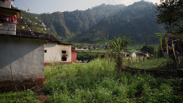 Lush green hillside homestead with colorful prayer flags on rooftop overlooking a misty terraced valley and distant Himalayan foothill village at golden hour