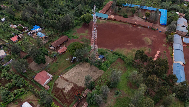 Aerial view of a telecommunications tower in a rural school compound with red soil, surrounded by trees, agricultural plots, and buildings with blue roofs in Gatundu, Kiambu County, Kenya.