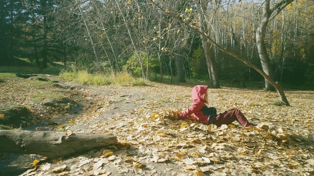 Boy lying in golden autumn leaves making a leaf angel then gathering them