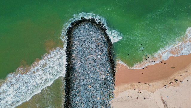 Aerial view of Tema Harbour breakwater with stone textures extending into the turquoise sea, sandy beach, and people walking along the shore in Tema, Greater Accra, Ghana.