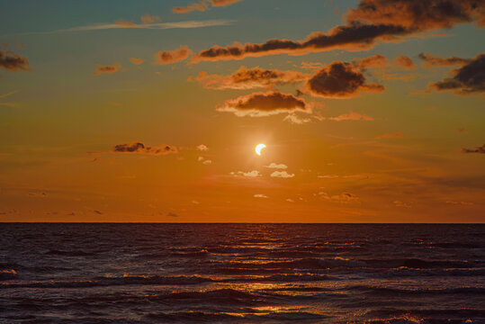 Partial solar eclipse glowing above the ocean at sunset, with orange clouds and shimmering reflections across the dark waves.