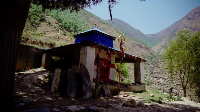 Rustic Hindu shrine with blue tin roof and golden trident overlooking rocky mountain riverbed in remote Himalayan Nepal valley