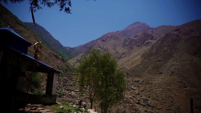 Golden trident of rural shrine framing rugged rocky Himalayan mountain valley with distant hikers walking through dry riverbed terrain