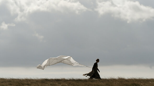 Kite builder carrying a finished geometric cellular kite across an open hillside under a brooding overcast sky, aftermath of a long construction session visible in fabric scraps