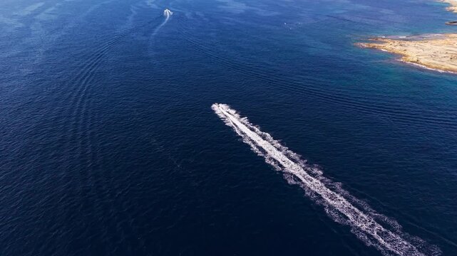 A jet ski races across the open blue sea off Malta, leaving a long white wake, chased directly overhead by a forward-moving drone toward another vessel ahead.