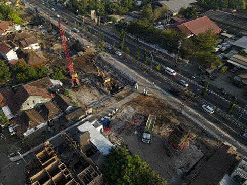 Aerial view of the construction of the Solo-Yogyakarta Toll Road with a red crane and excavators operating near a busy highway in Kecamatan Depok, Daerah Istimewa Yogya, Indonesia.