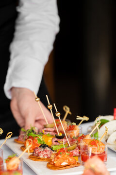 A waiter serving a platter of elegant gourmet appetizers, including tartare canap&eacute;s, shrimp shooters, and skewered hors d'oeuvres at a catered event.