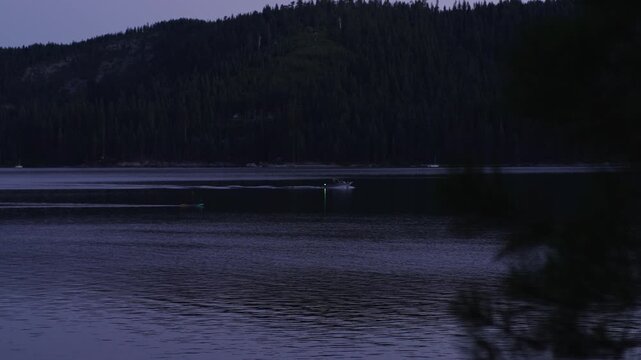 Paddleboarder and motorboat cross dark mountain lake at twilight, leaving separate wakes across calm water beneath a forested shoreline and dim evening sky