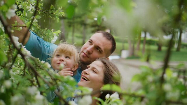 Family smiles through blooming tree branches. Mom, dad, and child enjoy spring outdoors. They look up, happy, curious. Bright green leaves frame their joyful faces. Perfect for nature, family