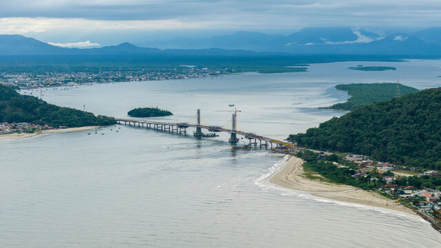 Aerial view of the construction of the Ponte de Guaratuba bridge spanning the bay with surrounding lush green hills, sandy beaches, and calm waters in Guaratuba, Parana, Brazil.