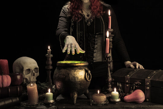 Witch casts spell, cauldron, boiling magic potion, candles, skull. Woman witchcrafting, magician girl with red hair, dressed in Halloween attire