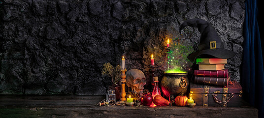 Halloween background. Dark witch cauldron with a magic witch's potion, books, chest, conical hat. Witchcraft paraphernalia and accessories among candles on a table in a room or cave with stone walls. © TSViPhoto