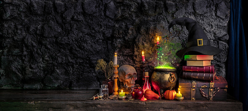 Halloween background. Dark witch cauldron with a magic witch's potion, books, chest, conical hat. Witchcraft paraphernalia and accessories among candles on a table in a room or cave with stone walls.