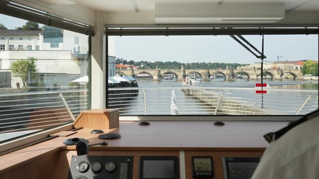 Ship wheelhouse view navigating the Vltava river toward Charles Bridge in Prague. The wooden captain dashboard overlooks the urban waterway and historical city architecture on a sunny summer day.