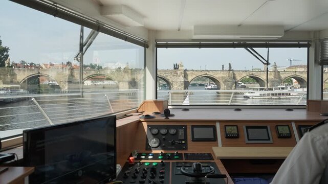 Ship captain maneuvers vessel toward Charles Bridge on Vltava river in Prague. Modern bridge control console guides tourist cruise boat. Professional nautical navigation in summer Europe.