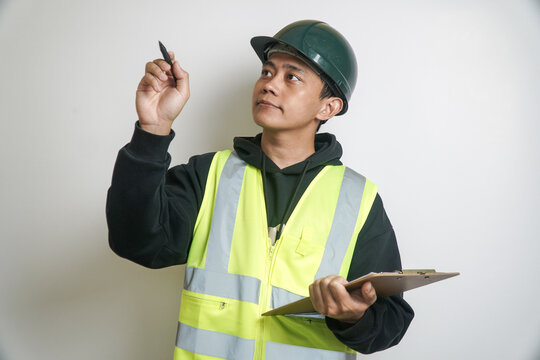 Young Asian Male Engineer in Safety Vest and Hard Hat Inspecting, Looking Up While Holding a Pen and Clipboard