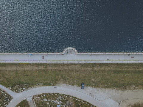Aerial view of Lac des Bouillouses dam with a stone wall, calm blue water, grassy embankment, and a single car parked on the gravel path in Pyrenees-Orientales, Occitanie, France.